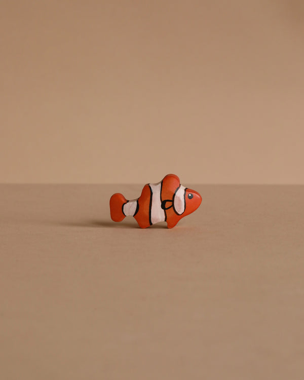 A Handmade Holzwald Clownfish, featuring orange and white colors with black stripes, sits on a plain beige surface with a matching background—an adorable addition to any Holzwald wooden toy collection.