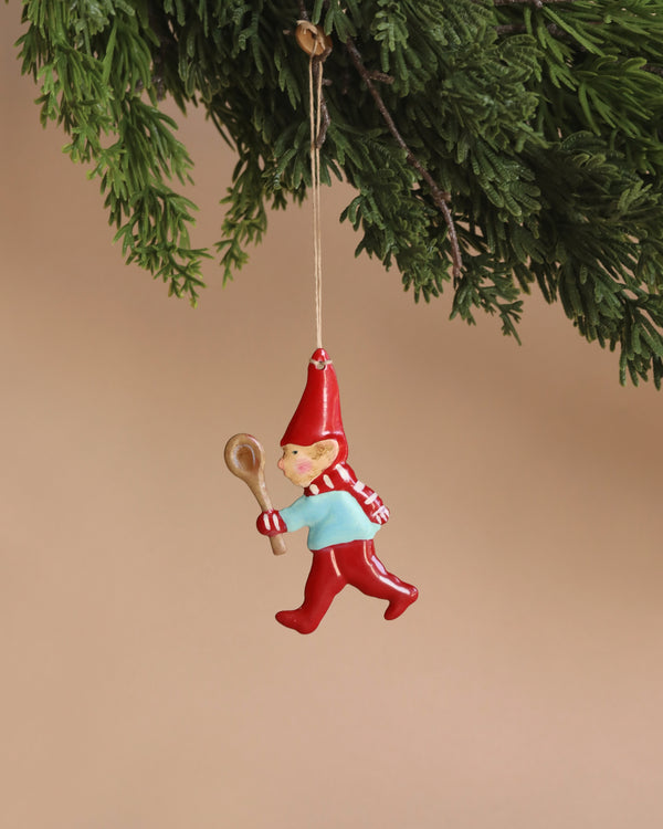 Hanging from a green Christmas tree branch against a beige background, the Maileg Metal Ornament, Pixy Spoon features a jolly elf adorned in red and blue, with a pointy hat and striped scarf. It showcases intricate handpainted details as it holds a wooden spoon.