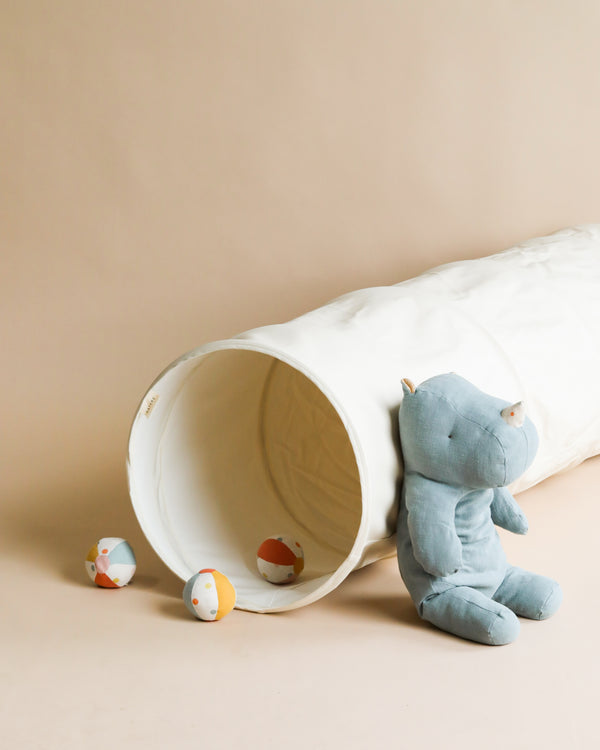 A blue plush hippopotamus toy sits beside a Gathre Vegan Leather Play Tunnel on a beige background, with three small, colorful balls scattered around.