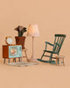 The Maileg Family Room Set features a green rocking chair, small stool, lamp, TV with a butterfly on the screen, clock, potted plant, and woven rug—all perfectly designed for your dollhouse and beautifully set against a peach background.