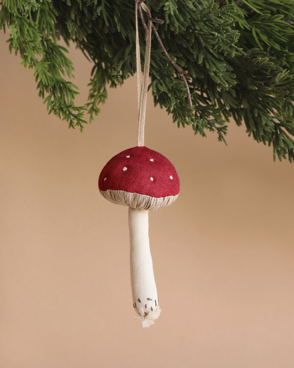 The Maileg Mushroom Ornament - Red is a delightful addition to your Christmas home decoration. Featuring a charming red and white mushroom design, it hangs gracefully from a green pine branch against a beige background, capturing the essence of festive elegance.