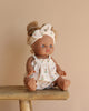 The Minikane Baby Doll (13") - Louise sits on a wooden stool against a neutral backdrop, dressed in a patterned outfit and matching headband. Its blue eyes and light brown hair in a bun complement its light brown skin and anatomically correct design.