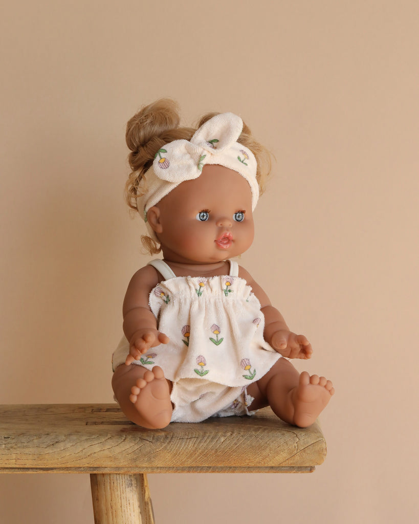 The Minikane Baby Doll (13") - Louise sits on a wooden stool against a neutral backdrop, dressed in a patterned outfit and matching headband. Its blue eyes and light brown hair in a bun complement its light brown skin and anatomically correct design.