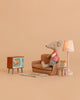 A Maileg Mouse in a striped shirt relaxes on a small couch, surrounded by the Maileg Cozy TV Time Set, which includes charming dollhouse furniture and accessories—a lamp, alarm clock, and cozy rug—all arranged against a beige background as it enjoys watching TV.