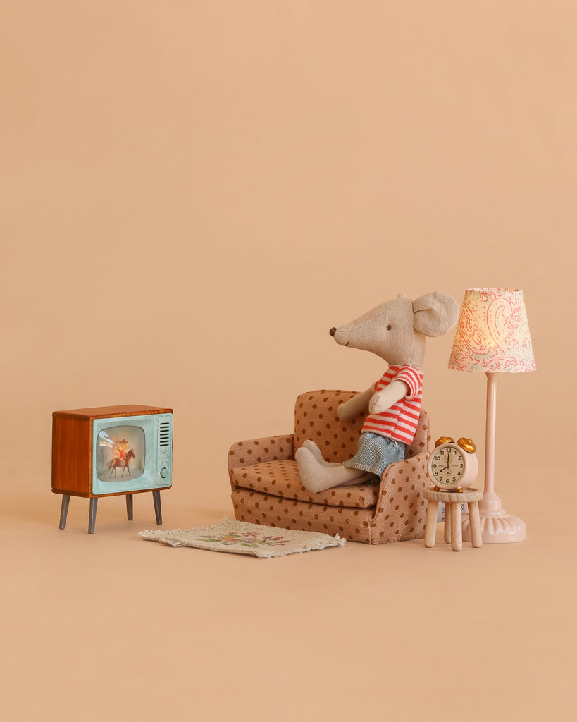 A Maileg Mouse in a striped shirt relaxes on a small couch, surrounded by the Maileg Cozy TV Time Set, which includes charming dollhouse furniture and accessories—a lamp, alarm clock, and cozy rug—all arranged against a beige background as it enjoys watching TV.