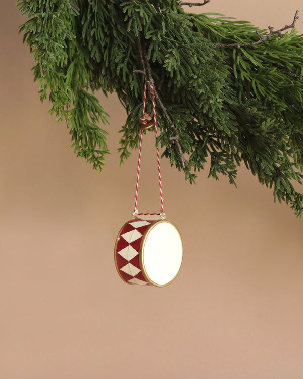 A large Maileg Metal Ornament in the shape of a drum, featuring red and white diamond patterns, gracefully hangs from a tree branch adorned with lush green needles against a beige background, highlighting its exquisite handpainted details.