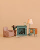 The Maileg Living Room Set includes charming Maileg mouse furniture—a pink armchair, potted plant, and a blue miniature Maileg fireplace—accented with a gold clock, patterned lamp, and vintage-style TV. Beautifully arranged on a beige background, these Maileg dollhouse accessories create the perfect miniature living room scene.