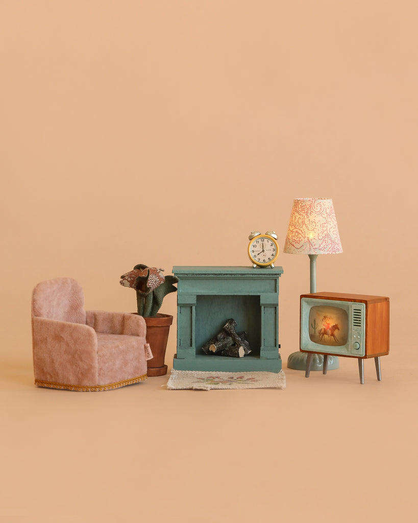 The Maileg Living Room Set includes charming Maileg mouse furniture—a pink armchair, potted plant, and a blue miniature Maileg fireplace—accented with a gold clock, patterned lamp, and vintage-style TV. Beautifully arranged on a beige background, these Maileg dollhouse accessories create the perfect miniature living room scene.