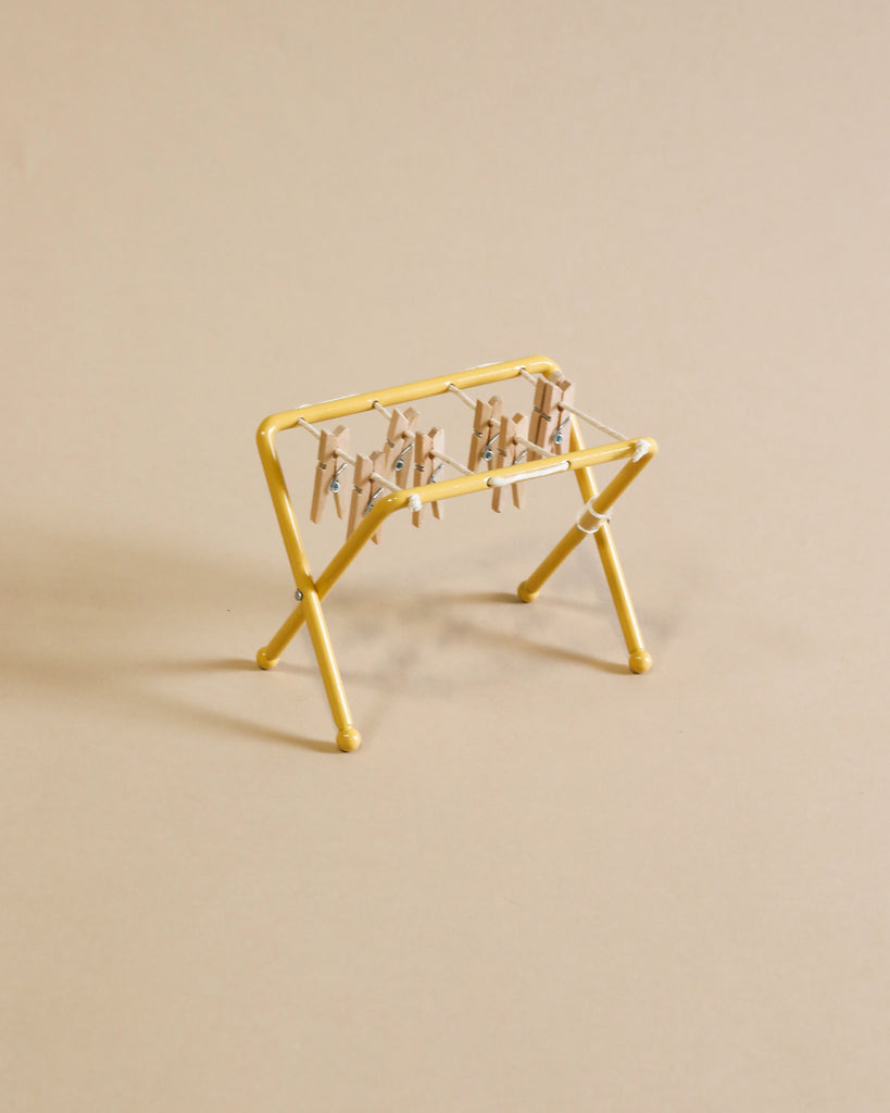 The Maileg Laundry Room Set features a small yellow drying rack with miniature clothespins, displayed on a beige surface for a simple, minimalistic look.