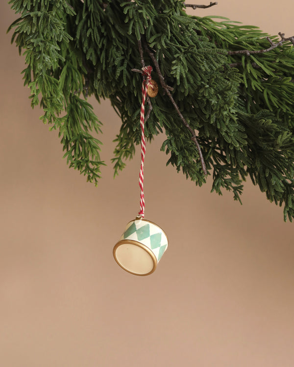 A Maileg Metal Ornament, Small Drum - Mint, adorned with hand-painted green and white diamond patterns, dangles elegantly from a red and white striped string on a pine branch. Its delightful design pops against the plain light brown background.