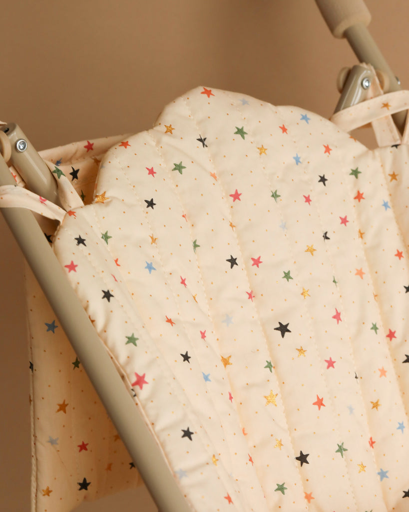 A close-up of a baby swing seat with a fabric cover featuring a beige background and a pattern of colorful stars in various sizes. The Konges Sloejd Doll Stroller - Multi Star frame, which resembles the sturdiness of a doll stroller with double wheels, is beige, and the seat padding is plush and quilted for comfort.