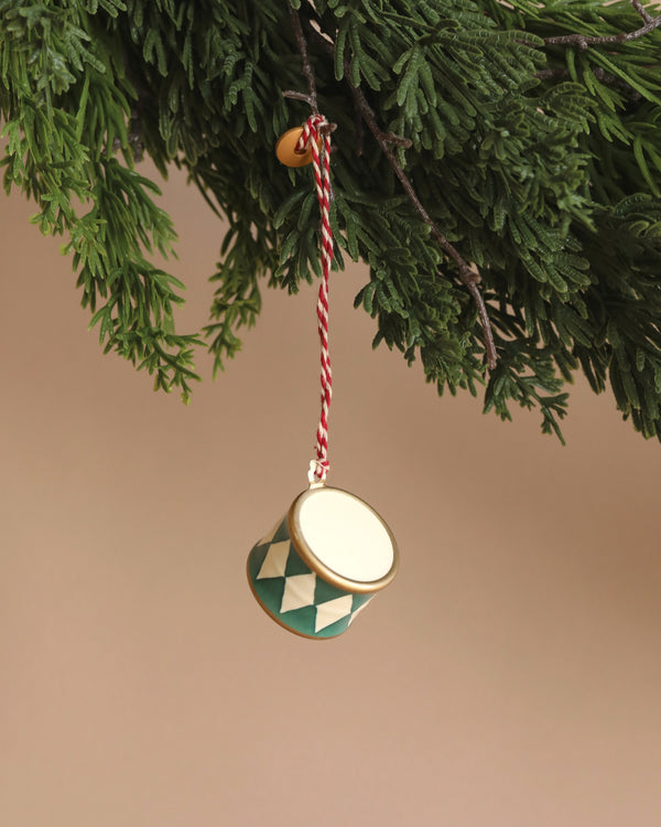 A Maileg Metal Ornament, Small Drum - Dark Green, featuring festive hand-painted green and white diamond patterns, hangs from a red and white string on a Christmas tree branch against a neutral background, complementing the charming collection of Maileg ornaments.