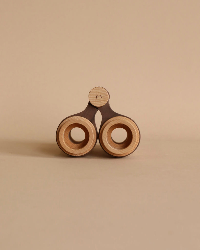 A minimalist image of a unique, solid beech wood-handled pair of Wooden Binoculars For Pretend Play against a neutral beige background. The design features a distinctive twin circular handle.