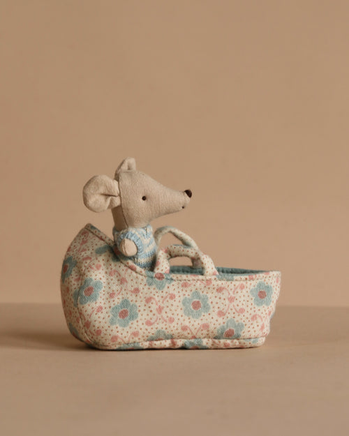 The Maileg Baby Mouse in Carry Cot - Blue features a tiny mouse in a knitted suit resting in a floral fabric cot, set against a plain beige background.