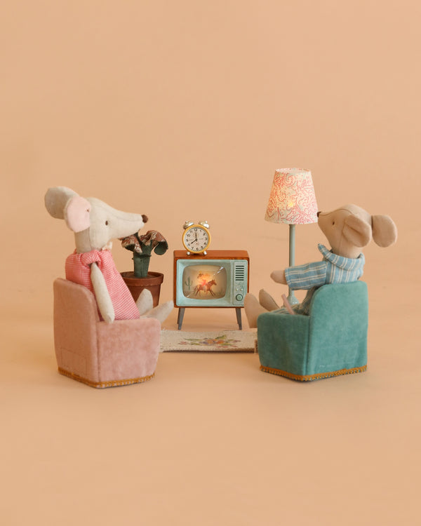 Two Maileg mice enjoy their time together in the Maileg Movie Night Set, relaxing in chairs as they watch TV. The cozy scene includes a lamp, plant, rug, and clock—charming Maileg dollhouse accessories—all set against a soft peach background.