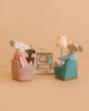 Two Maileg mice enjoy their time together in the Maileg Movie Night Set, relaxing in chairs as they watch TV. The cozy scene includes a lamp, plant, rug, and clock—charming Maileg dollhouse accessories—all set against a soft peach background.