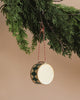 A Maileg Metal Ornament, Large Drum in dark green with a handpainted diamond pattern of green and gold hangs elegantly from a vibrant evergreen tree. The ornament is suspended by a charming red and white string, which stands out against the soft, neutral background.