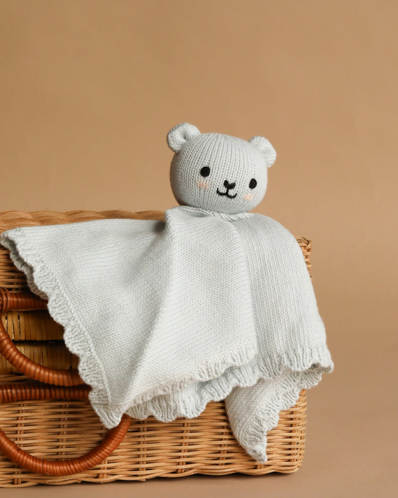 The Cuddle + Kind Organic Cotton Baby Bear Lovey (Baby Blue), a soft fair trade lovey with a knitted bear head and blanket body, is displayed over a woven wicker basket against a light brown background—perfect as a cozy baby gift.