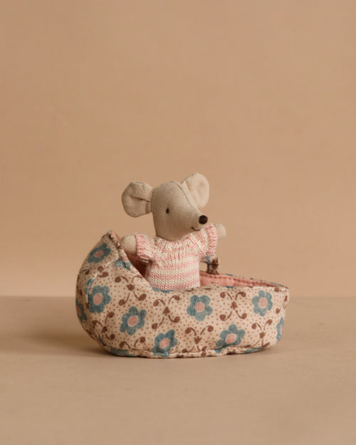 The Maileg Baby Mouse in Carry Cot - Rose features a small gray mouse in a pink sweater, resting inside a floral-patterned fabric carry cot, set against a beige background.
