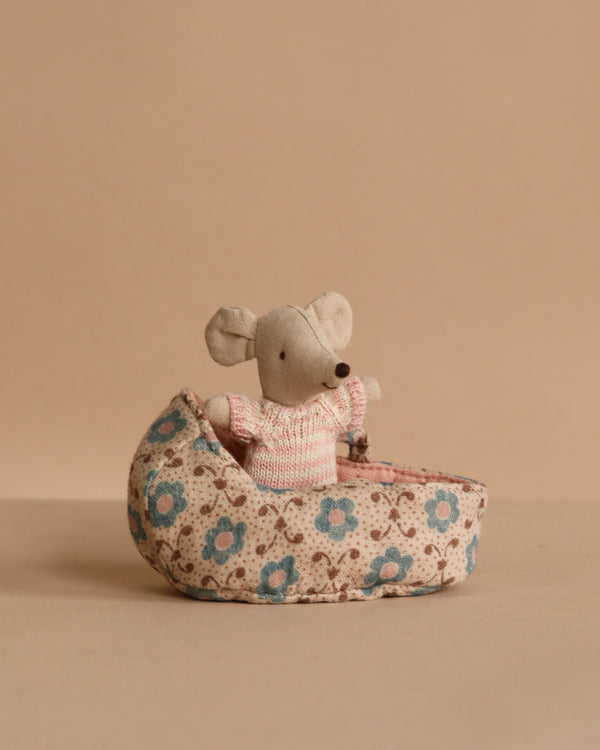 The Maileg Baby Mouse in Carry Cot - Rose features a small gray mouse in a pink sweater, resting inside a floral-patterned fabric carry cot, set against a beige background.