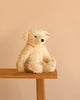 A Steiff Luca Teddy Bear, 12", with black eyes and fuzzy cream-colored fur sits on the edge of a wooden bench against a soft beige background, its signature "Button in Ear" marking hinting at its authentic Steiff origins.
