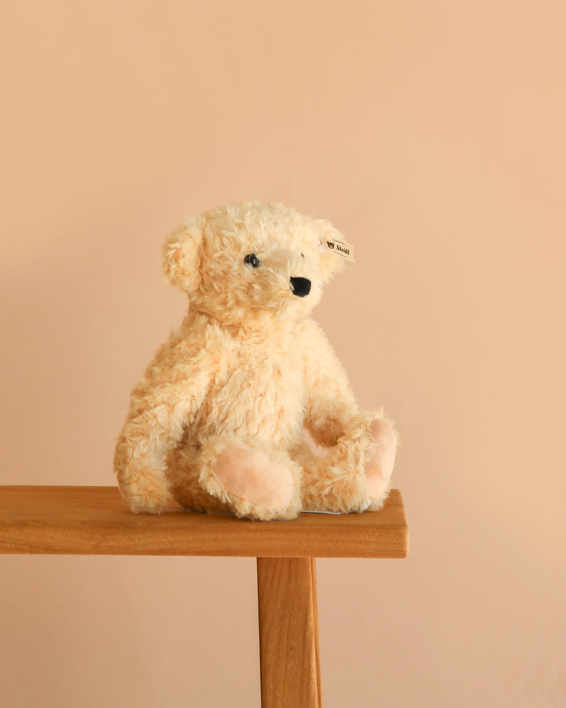 A Steiff Luca Teddy Bear, 12", with black eyes and fuzzy cream-colored fur sits on the edge of a wooden bench against a soft beige background, its signature "Button in Ear" marking hinting at its authentic Steiff origins.