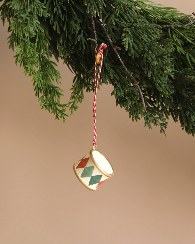 This delightful Maileg Metal Ornament, Small Drum - Classic Green is adorned with red and green diamond patterns and intricate handpainted details. It hangs from a green pine branch by a red and white string, beautifully contrasted against a neutral beige background.