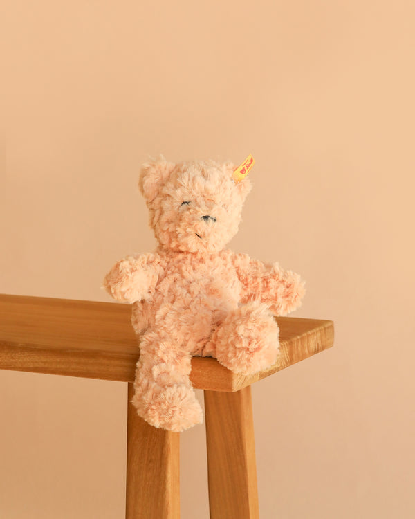 An 8" Steiff Baby Jimmy Teddy Bear, featuring soft, light peach plush fur, the classic Steiff Button in Ear, and a yellow tag, sits on the corner of a wooden bench against a beige background.