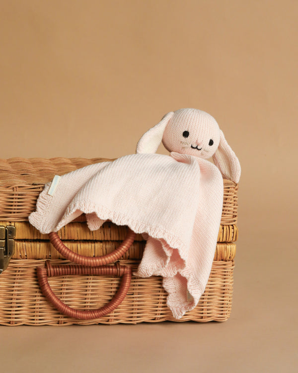 The Cuddle + Kind Organic Cotton Baby Bunny Lovey (Blush), a soft hand-knit pink bunny with floppy ears, rests on a closed wicker basket with two handles against a plain beige background.