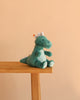 A Steiff Joshi Baby T-Rex Dinosaur in teal plush, featuring white spikes, sits on the edge of a light wooden table against a plain peach background—ready to become your child’s cuddly companion from the prehistoric past.