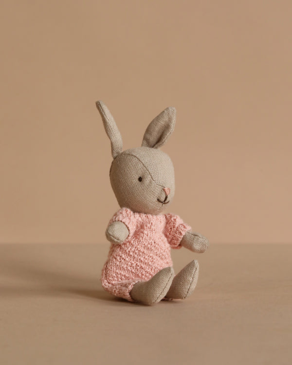 The Maileg Rabbit Micro - Sand wears a rose knitted suit and sits upright, featuring soft beige fur and long ears—ideal as a Maileg accessory or for a miniature dollhouse, photographed against a plain beige background.