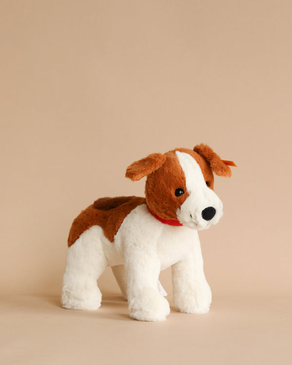 A Steiff Snuffy Dog Plush Puppy, 11" with white and brown patches and a red collar stands against a plain beige background. The machine washable plush is designed to look like a small, friendly puppy.