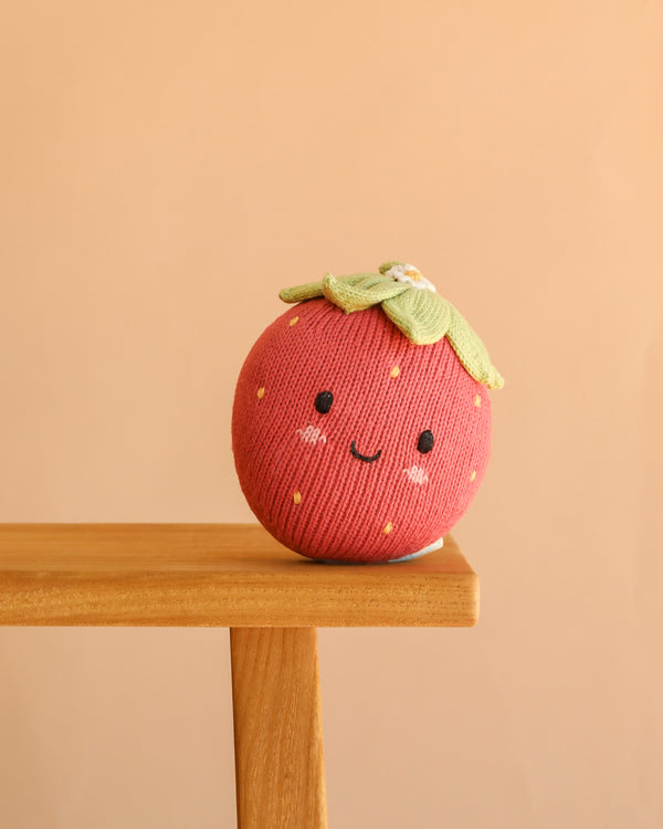 The Cuddle + Kind Baby Strawberry, a charming round plush with a smiling face, is handcrafted from heirloom-quality Peruvian cotton yarn and rests on the edge of a wooden surface against a peach-colored background.