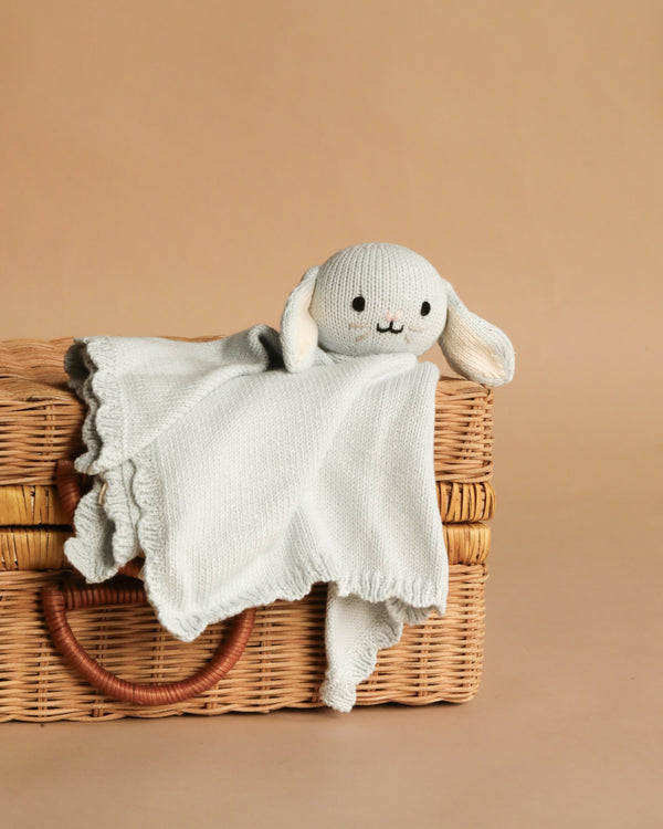 The Cuddle + Kind Organic Cotton Baby Bunny Lovey (Baby Blue) with floppy ears and a sweet smile drapes over a wicker basket, set against a plain tan background.