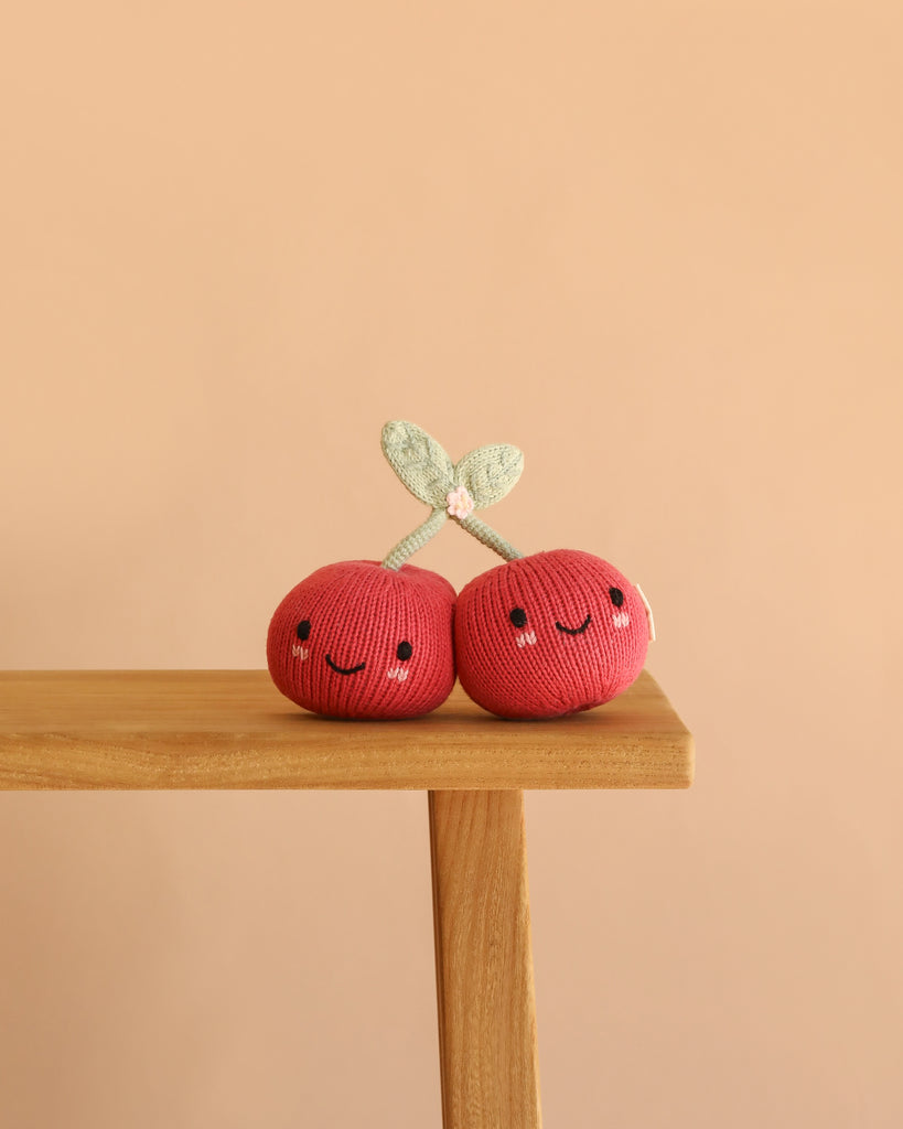 Two Cuddle + Kind Baby Cherry plush toys, each featuring smiling faces and green leaves and handcrafted from natural Peruvian cotton yarn, sit on the edge of a wooden table against a plain peach-colored background.