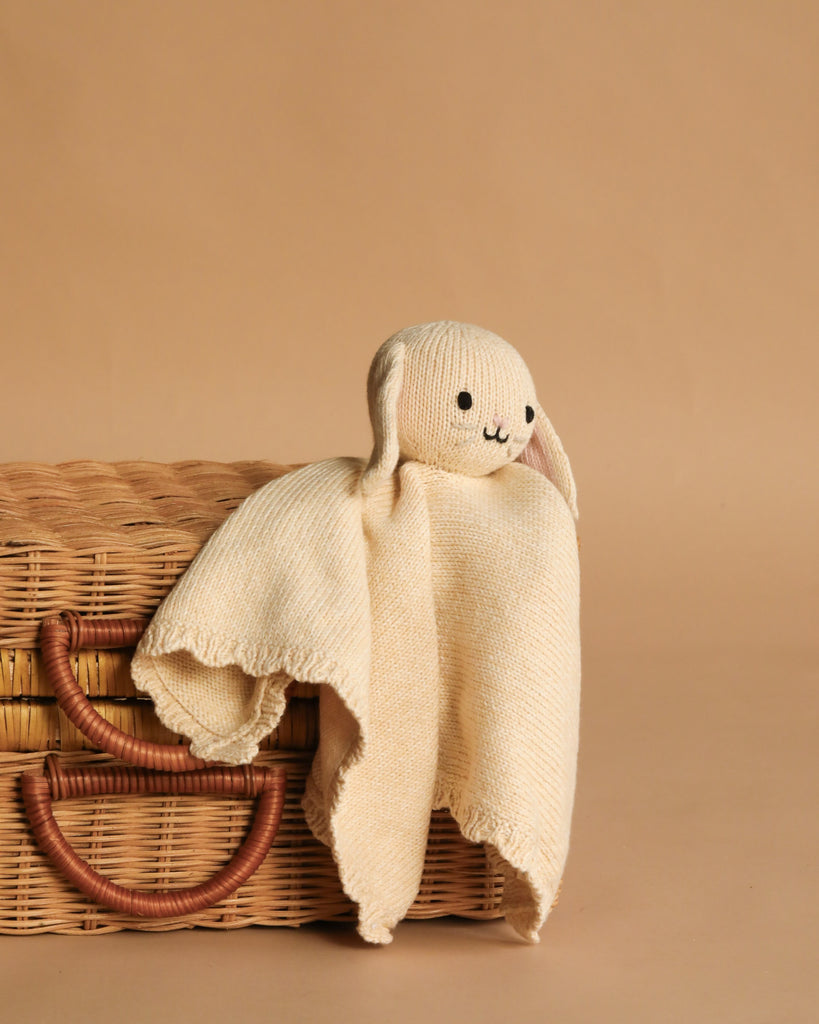 The Cuddle + Kind Organic Cotton Baby Bunny Lovey (Oatmeal), featuring stitched eyes, nose, and mouth, rests over a woven basket with leather handles against a beige background.