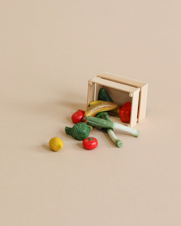 Miniature wooden crate tipped over with hand-painted Maileg Fruits & Veggies spilling out on a plain beige background.
