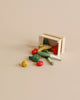 A small wooden crate tipped over, spilling out miniature vegetables—including broccoli, tomatoes, squash, leeks, and a lemon—onto a beige background: the perfect scene to complement the Maileg Family Celebration Set for creating charming displays or imaginative play with your Maileg mice in matchbox.