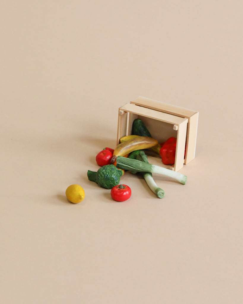 A small wooden crate tipped over, spilling out miniature vegetables—including broccoli, tomatoes, squash, leeks, and a lemon—onto a beige background: the perfect scene to complement the Maileg Family Celebration Set for creating charming displays or imaginative play with your Maileg mice in matchbox.