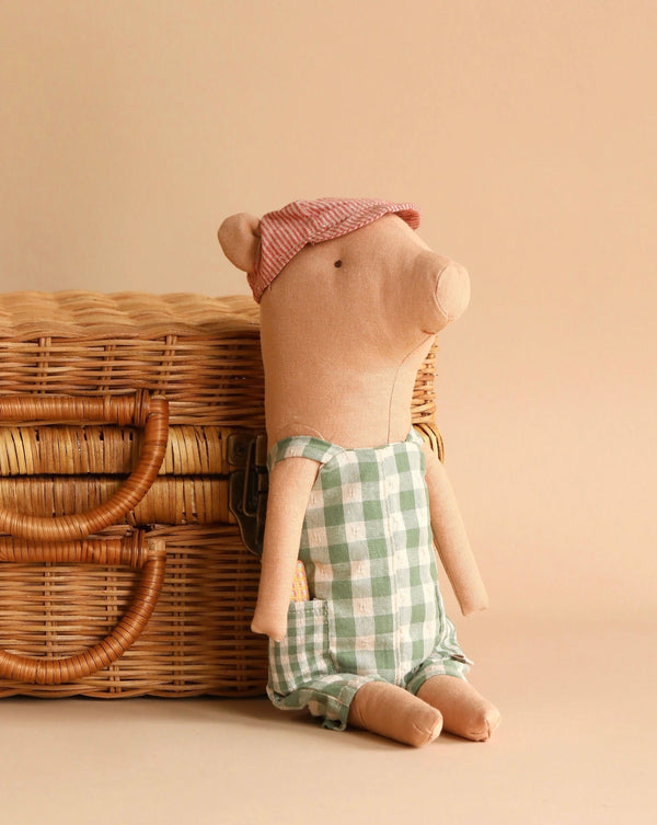 A Maileg Pig - Boy wearing a pink cap and green checkered romper sits beside a wicker basket. The background is beige, adding a soft, warm tone to the scene.