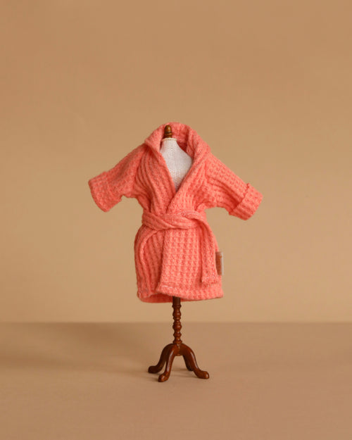 A small mannequin displays the Maileg Extra Clothing: Bathrobe - Coral, a textured pink cotton robe with a belt, set against a plain beige background.