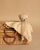 The Cuddle + Kind Organic Cotton Baby Bear Lovey (Honey) rests on a woven wicker basket with curved handles, set against a plain beige background.