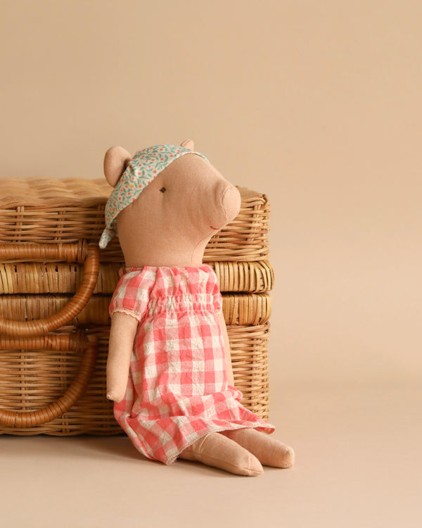 A Maileg Pig - Girl is sitting against a wicker basket, wearing a red and white checkered dress and a floral-patterned headband. The background is a warm beige color, complementing the natural tones of the wicker basket and the sweetest pig.