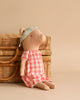 A Maileg Pig - Girl is sitting against a wicker basket, wearing a red and white checkered dress and a floral-patterned headband. The background is a warm beige color, complementing the natural tones of the wicker basket and the sweetest pig.