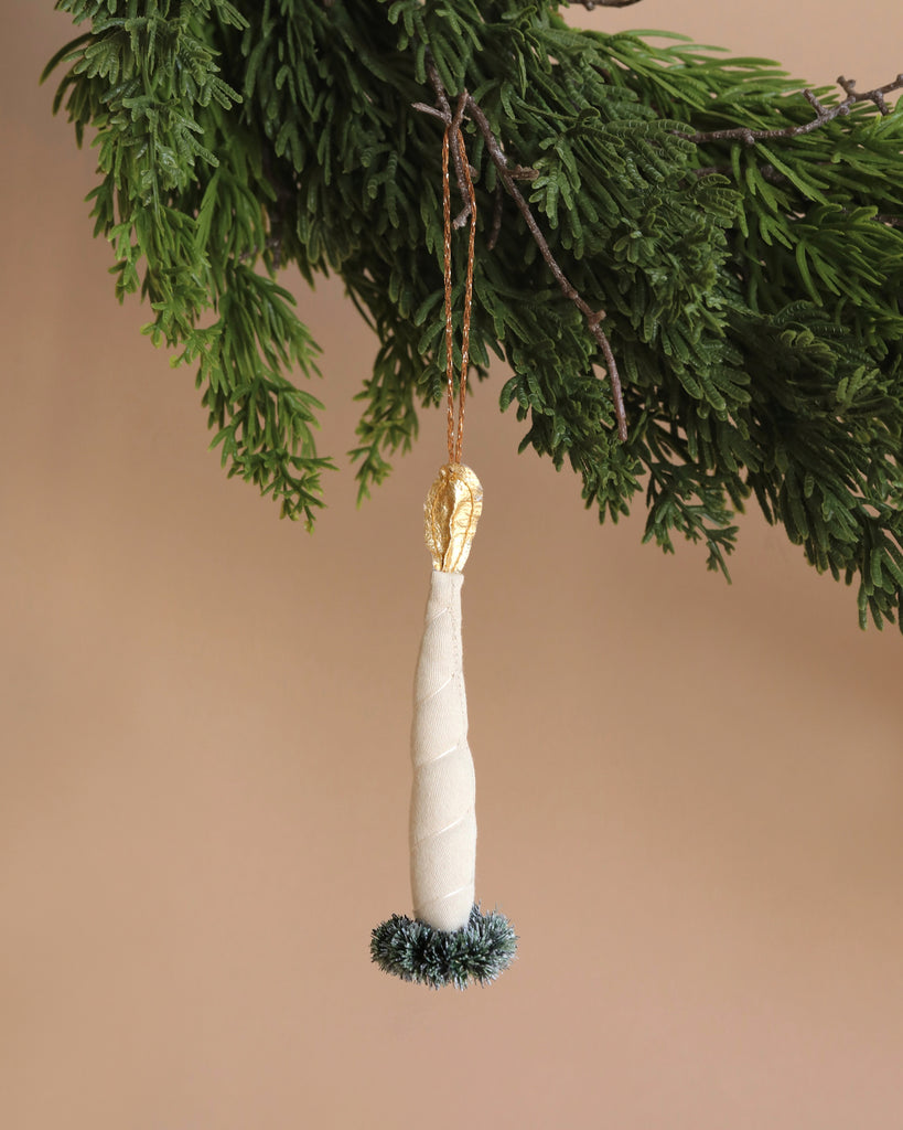 The Maileg Ornament - Candle Light, featuring a white and gold candle shape, hangs elegantly from a green branch. The base is decorated with green tinsel, adding to its festive charm. Placed against a neutral beige background, the ornament's intricate details are beautifully highlighted.