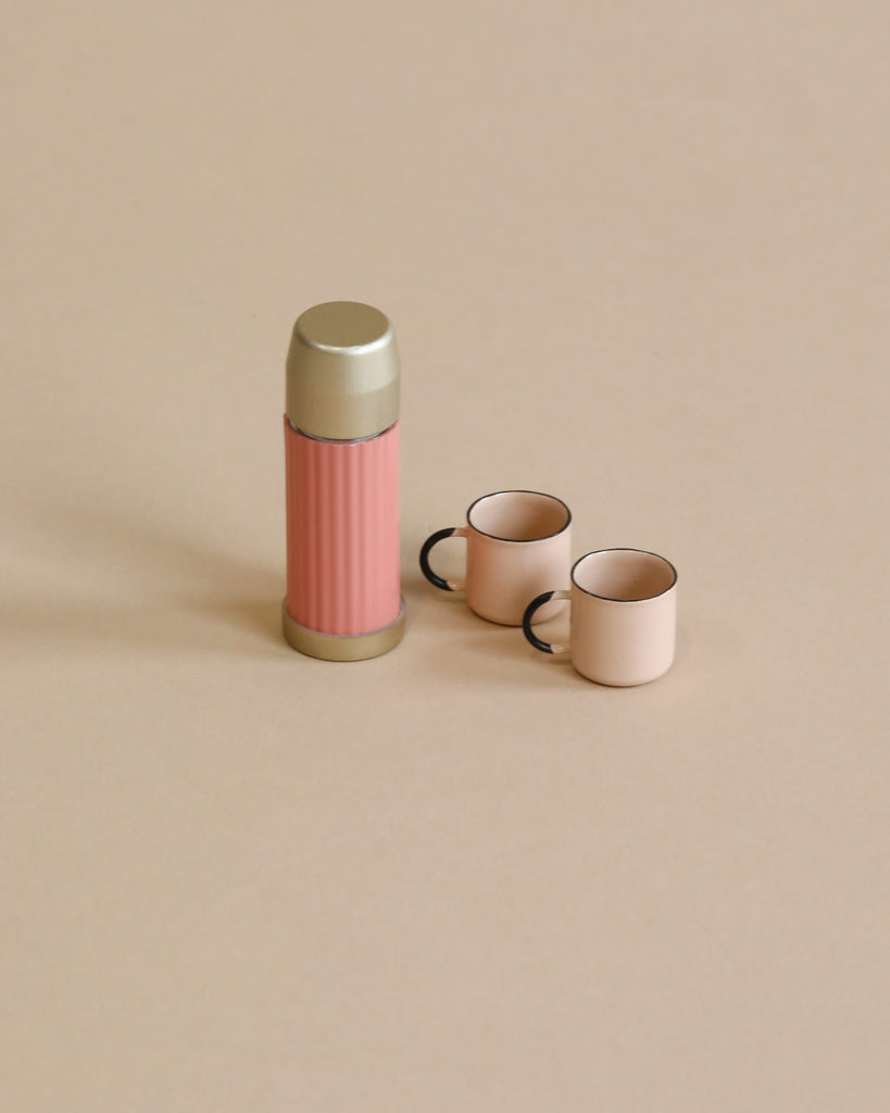 A Maileg Miniature Thermos And Cups with a grey lid next to two small pink cups with magnetic hands on a beige background.