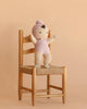 The Cuddle + Kind My First Baby (Lavender), a hand-knit doll dressed in a soft lavender outfit and bonnet and crafted from natural cotton yarn, sits on a wooden chair with a woven seat against a plain beige background.