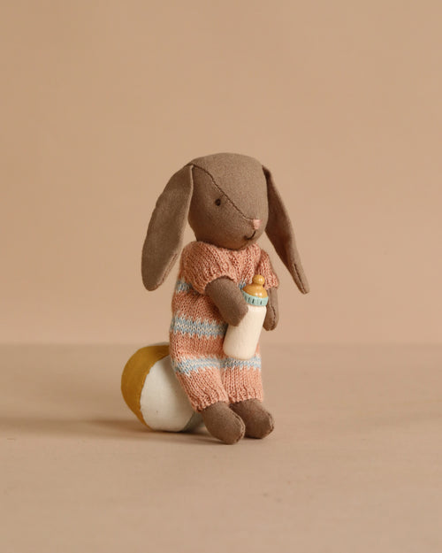 The Maileg Bunny With Accessories, Micro - Brown - Dusty Rose wears a peach and blue striped knitted outfit and sits on a yellow and white cushion holding a baby bottle—a charming collectible for any bunny lover.