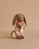 The Maileg Micro Bunny Bedroom Set features a plush brown bunny in a peach and blue striped sweater, sitting on a round yellow and white pillow with a small baby bottle, against a plain beige background.
