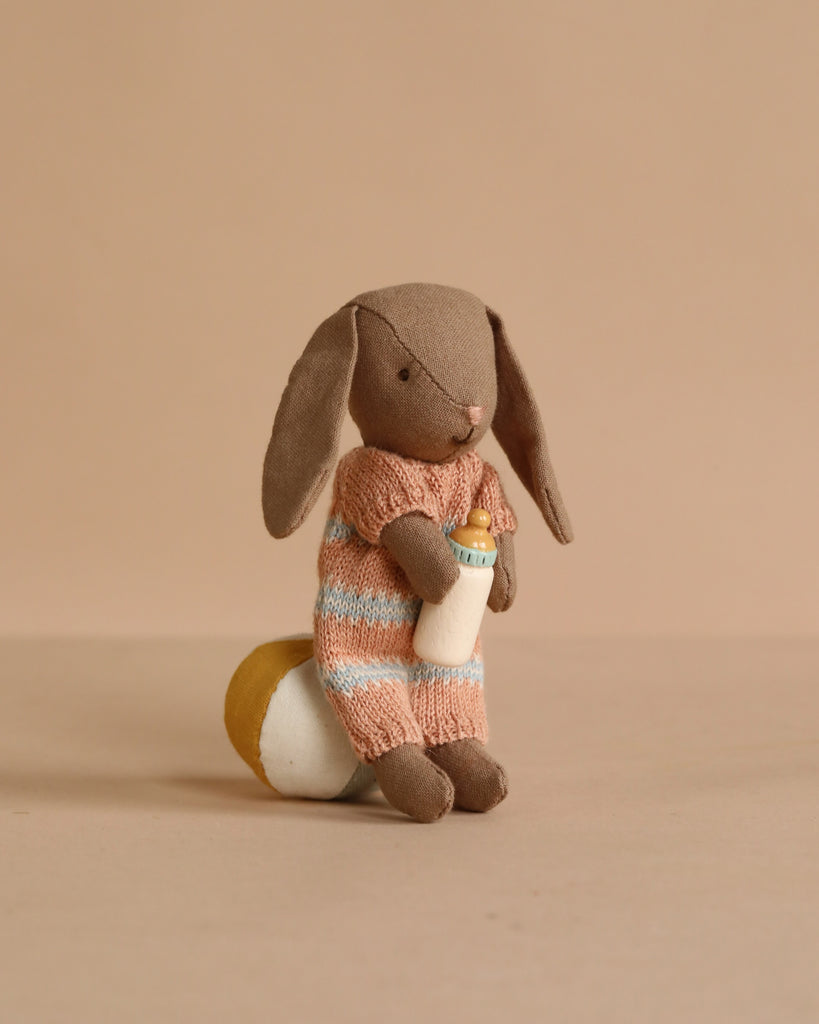 The Maileg Micro Bunny Bedroom Set features a plush brown bunny in a peach and blue striped sweater, sitting on a round yellow and white pillow with a small baby bottle, against a plain beige background.